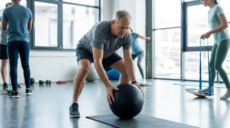 A fit older adult performing a high-velocity medicine ball training session in a modern rehabilitation and exercise studio under professional clinical supervision. Figure 1: Explosive power training in older adulthood. Moving moderate loads with high velocity targets Type II muscle fibers, directly addressing age-related declines in muscle power and rate of force development.